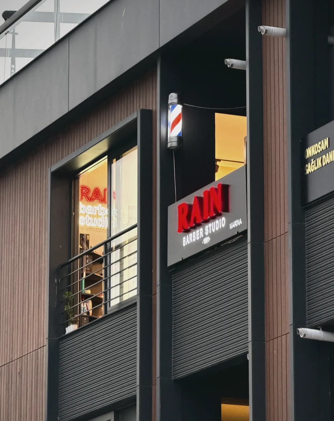 Rain Barber Studio'nun dış cephesi: ışıklı tabela, klasik barber direği ve camdan görünen neon